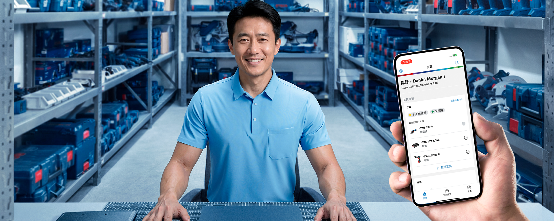 一个穿着浅蓝色衬衫的男人坐在桌子旁，桌子前是满是蓝色工具箱的架子。他微笑着，手里拿着一部显示应用程序的智能手机。环境光线良好，看起来很有条理。