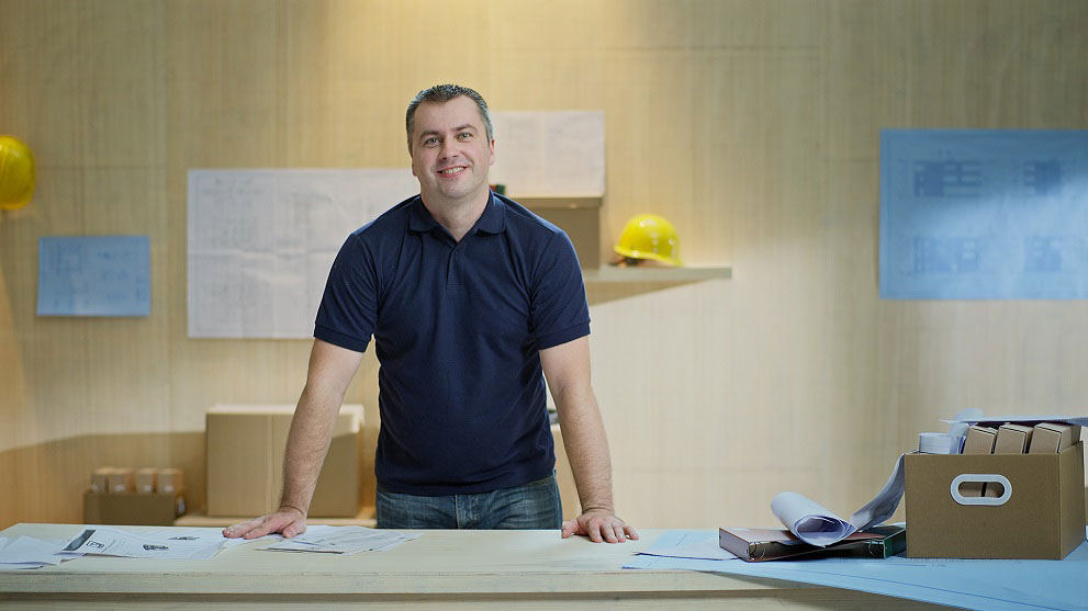 一个穿着蓝色衣服的微笑男人站在一张有建筑图纸和文件夹的桌子旁。背景中可以看到黄色的安全帽和图纸。
