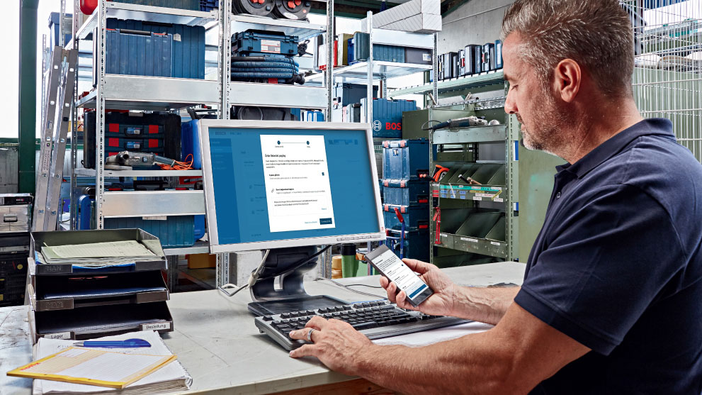 一个男人在电脑上工作，同时手里拿着一部智能手机。背景中有装满工具箱和材料的架子。